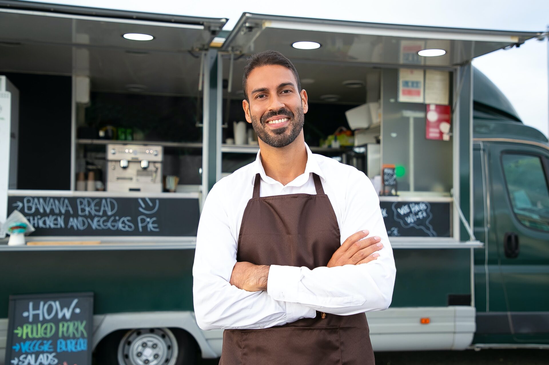 A man wearing an apron smiling in front of his small business food truck, posing with his arms crossed.